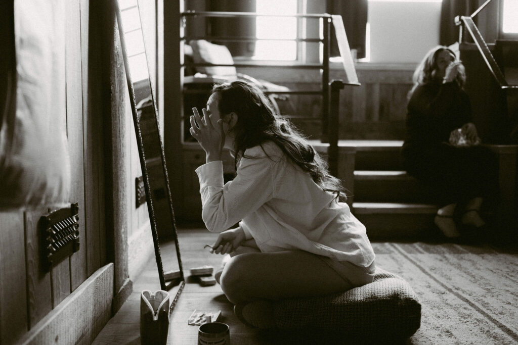 The bride applies makeup in soft morning light while sitting on the floor inside Timberline Lodge before her Silcox Hut wedding.