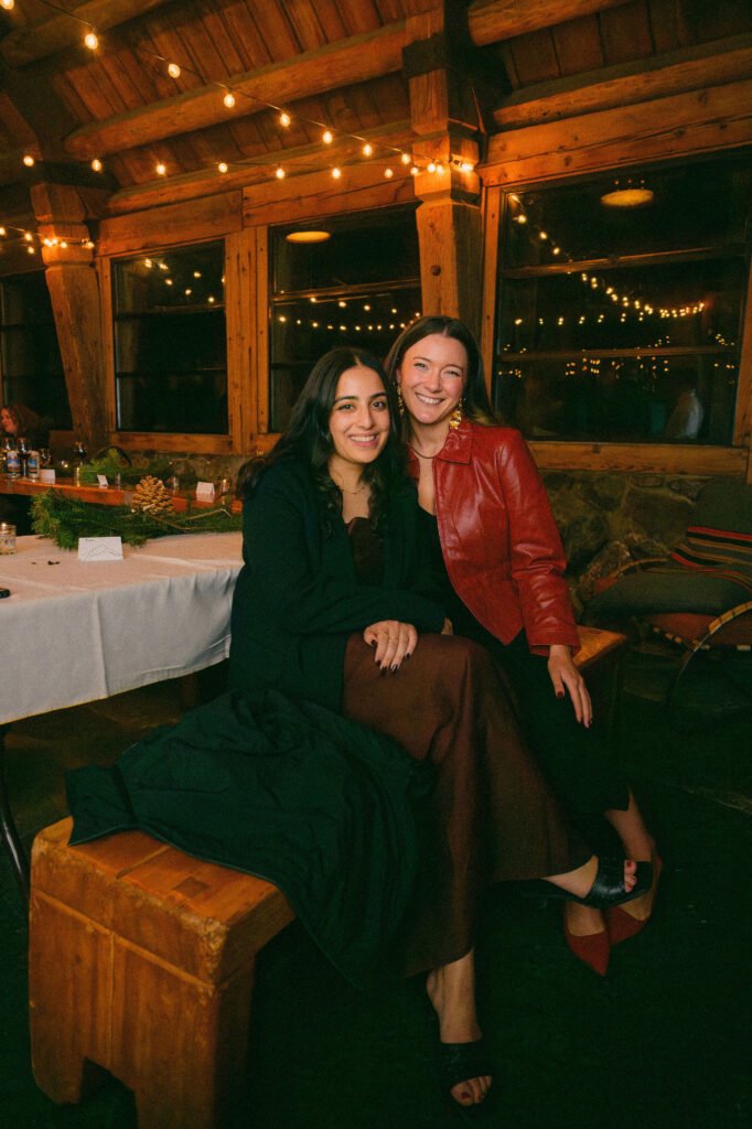 Two guests smile together inside Silcox Hut, seated beneath soft string lights during the cozy mountain wedding reception.