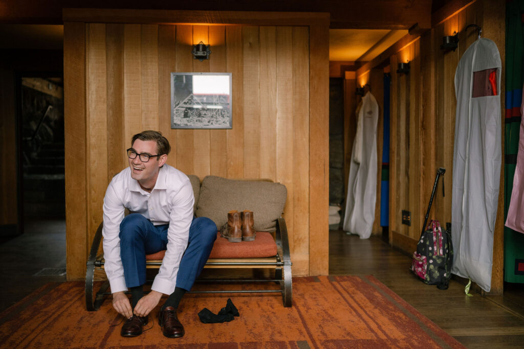 The groom ties his shoes while getting ready inside Silcox Hut, surrounded by warm wood paneling and vintage details.