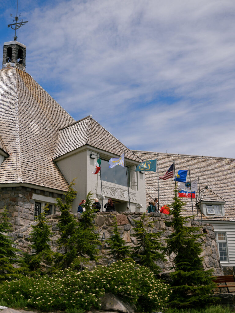 The historic stone exterior of Timberline Lodge with flags waving above lush summer greenery on Mount Hood.