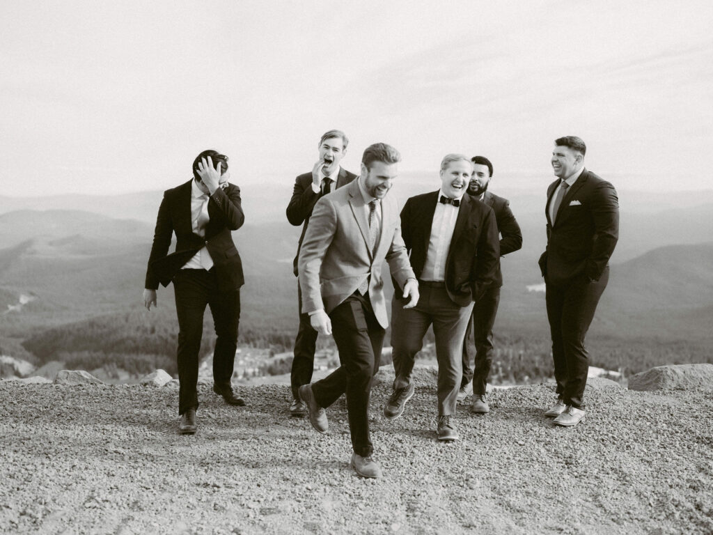 The groom and his groomsmen walk and laugh together on a windy ridge overlooking Mount Hood’s forested valleys.