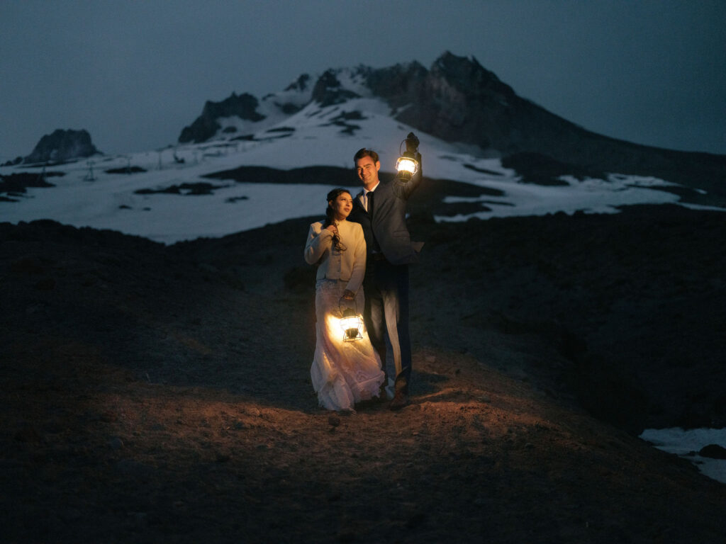 A bride and groom hold glowing lanterns at dusk on the snowy slopes of Mount Hood during their Silcox Hut wedding portraits.