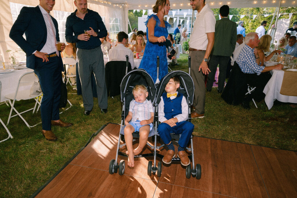 Two young boys asleep in side-by-side strollers on the dance floor during the wedding reception, surrounded by smiling guests.