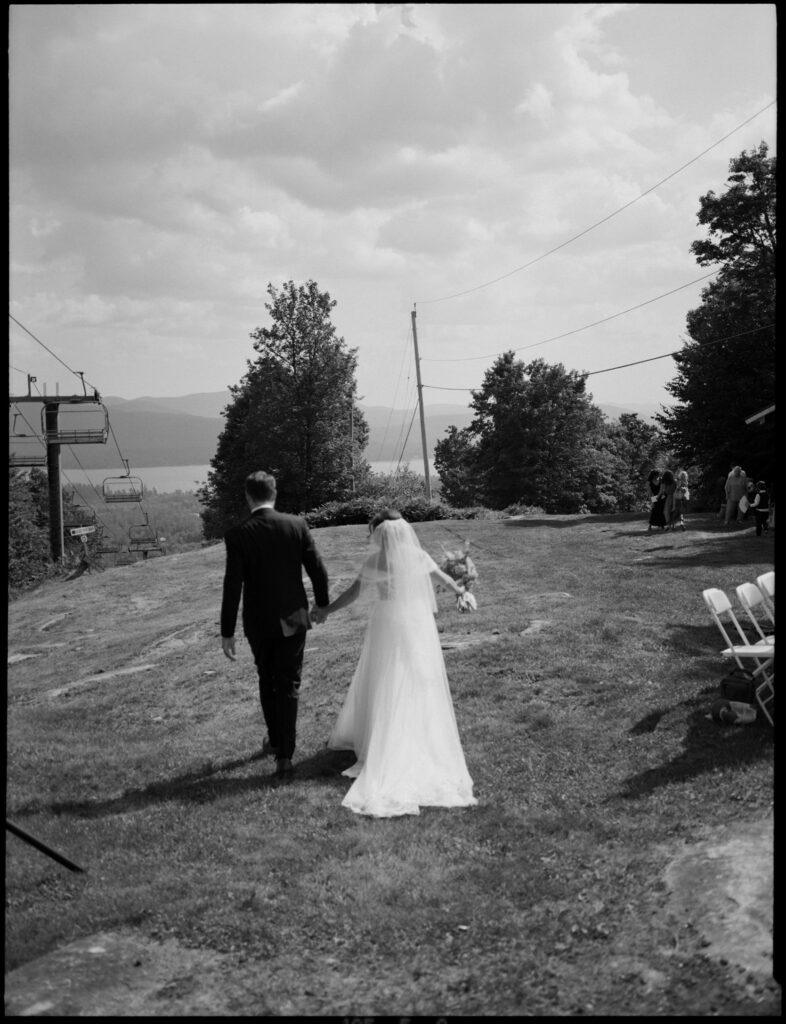 Black-and-white image of the bride and groom walking hand-in-hand across the hilltop at Oak Mountain, the lake and mountains visible in the distance.