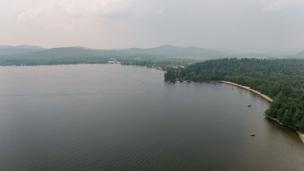 A wide aerial view of Lake Pleasant in Speculator, NY, surrounded by summer greenery and distant Adirondack mountains under a hazy sky.