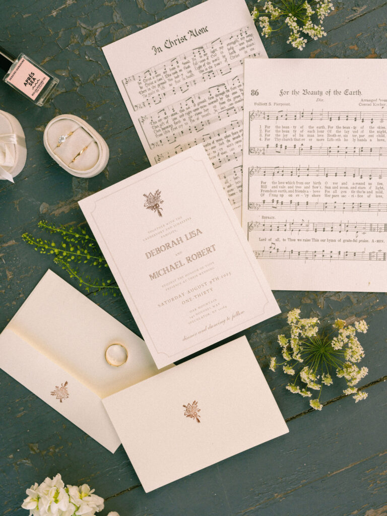 A styled flatlay of the wedding invitation suite, rings, perfume, and hymn sheets arranged with small white flowers on a rustic blue tabletop.