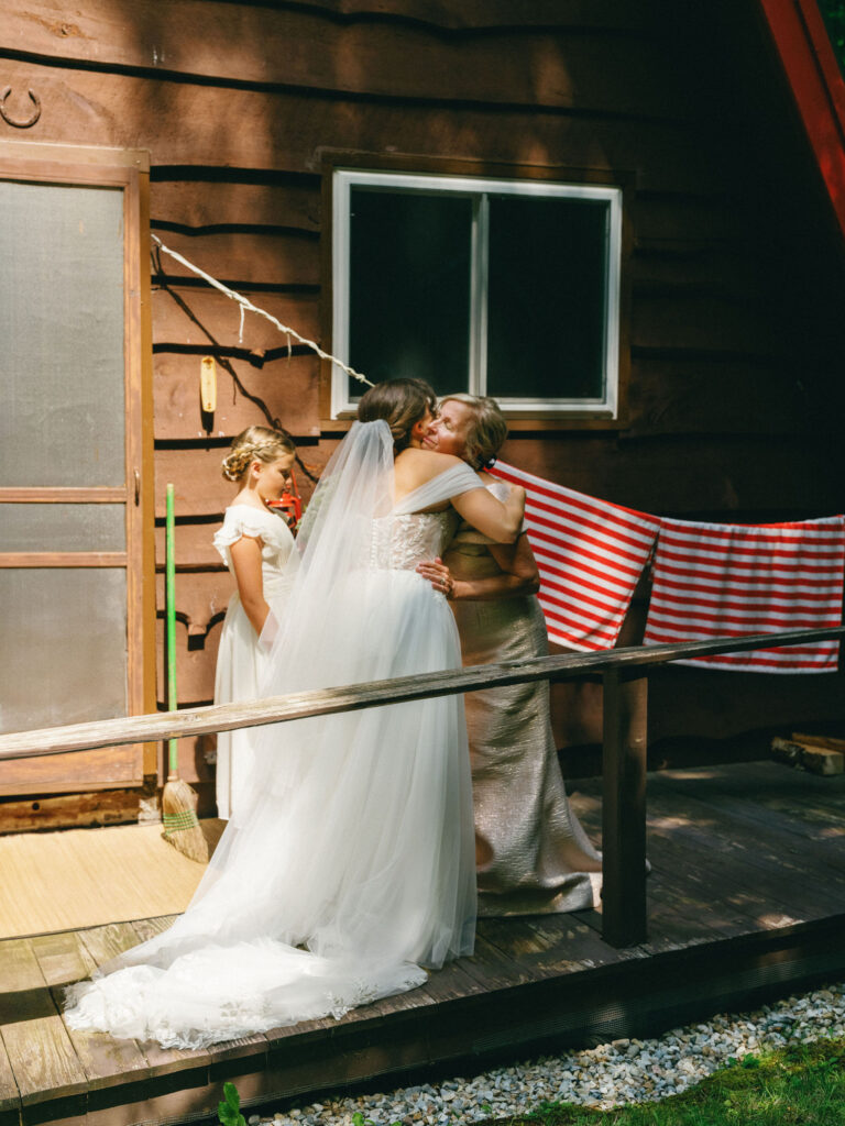 The bride embraces her mother on the porch of the family’s A-frame cabin in Speculator, NY, sunlight filtering across her veil and gown.