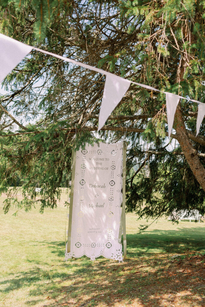 Hanging paper-cut welcome banner reading “Welcome to the Wedding of Deborah & Michael,” displayed under pine trees.