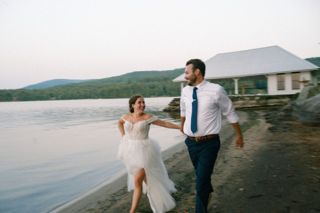 Bride and groom running hand-in-hand along the shoreline of Lake Pleasant at sunset during their Speculator, NY wedding.
