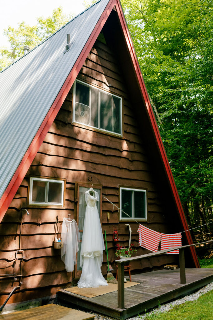 A wedding dress hangs on the porch of a rustic Adirondack A-frame cabin surrounded by summer greenery in Speculator, NY.