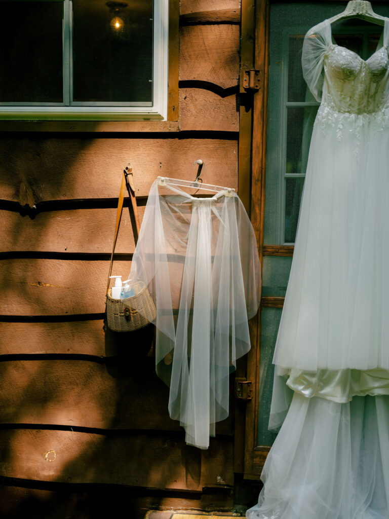 A wedding veil and gown hang on the exterior of the A-frame cabin, illuminated by soft patches of summer light.