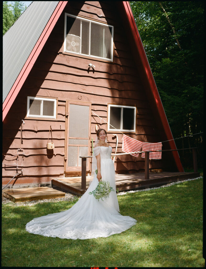 Film photograph of the bride standing on the grass before the family’s A-frame cabin, her long train spread behind her in the afternoon sun.