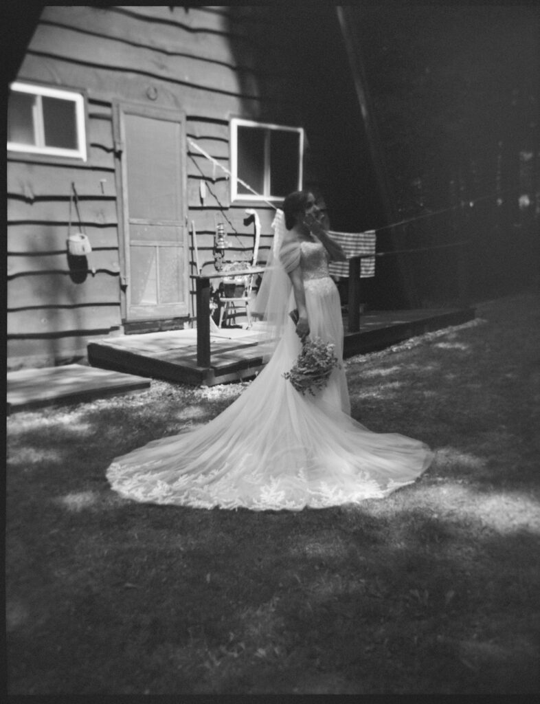 Black-and-white portrait of the bride standing in the yard near the cabin, holding her bouquet with Adirondack woods behind her.
