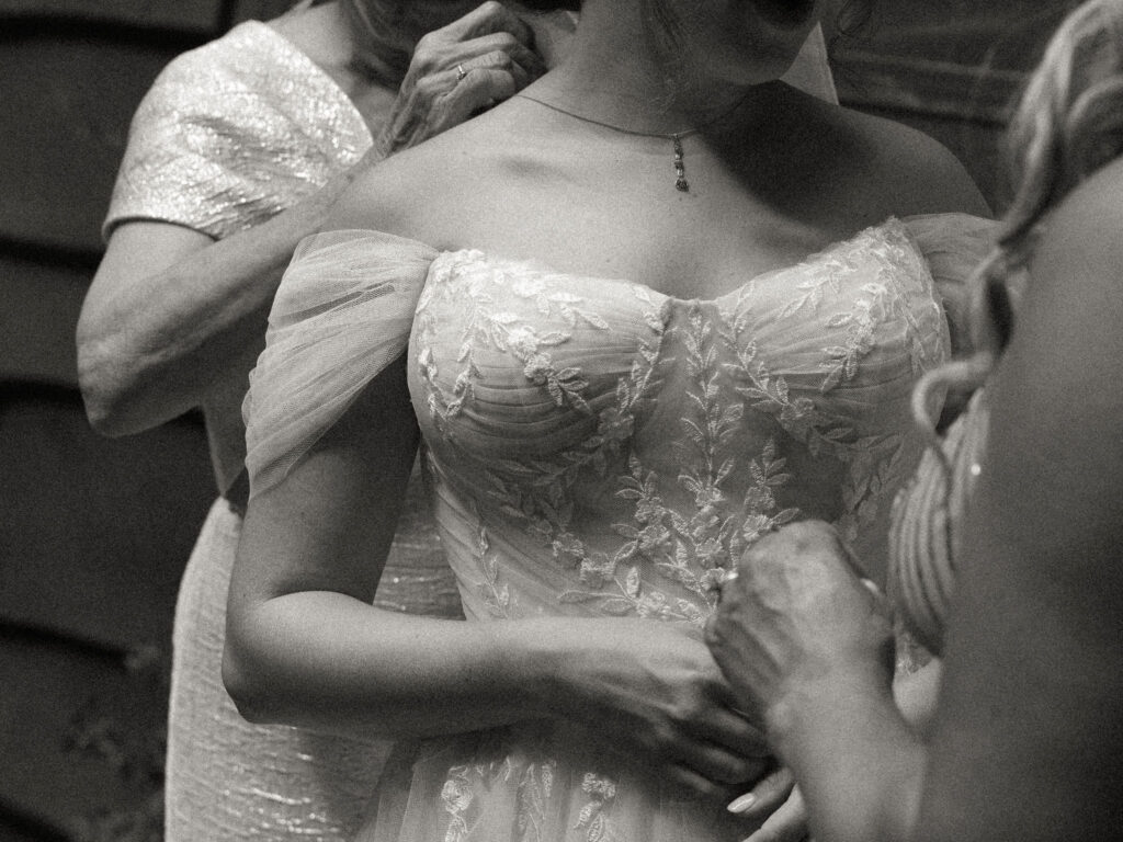 A close-up of the bride’s embroidered bodice as family members fasten her gown on the cabin porch, captured in black and white.