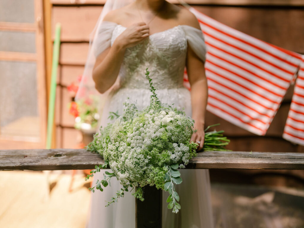 A lush white-and-green bridal bouquet rests on the wooden porch railing of the A-frame cabin as the bride stands behind it in her off-the-shoulder gown.