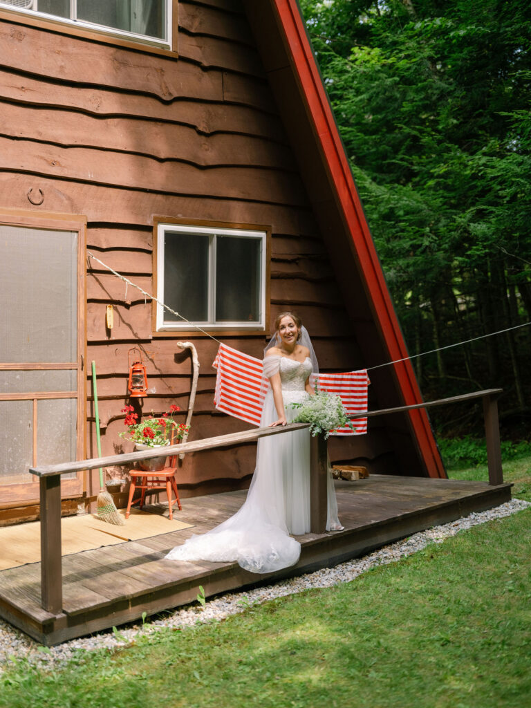 The bride stands on the porch of the A-frame cabin, leaning gently on the railing with her bouquet as sunlight filters through the surrounding trees.