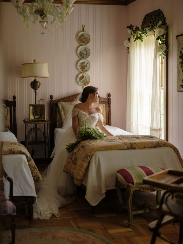The bride sits on the edge of a vintage bed, bouquet in hand, gazing toward the window as warm afternoon light fills the room.