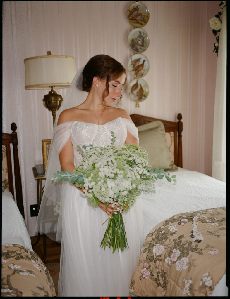 Film photograph of the bride standing between twin beds in a vintage bedroom, holding a full white-and-green bouquet and looking softly to the side.