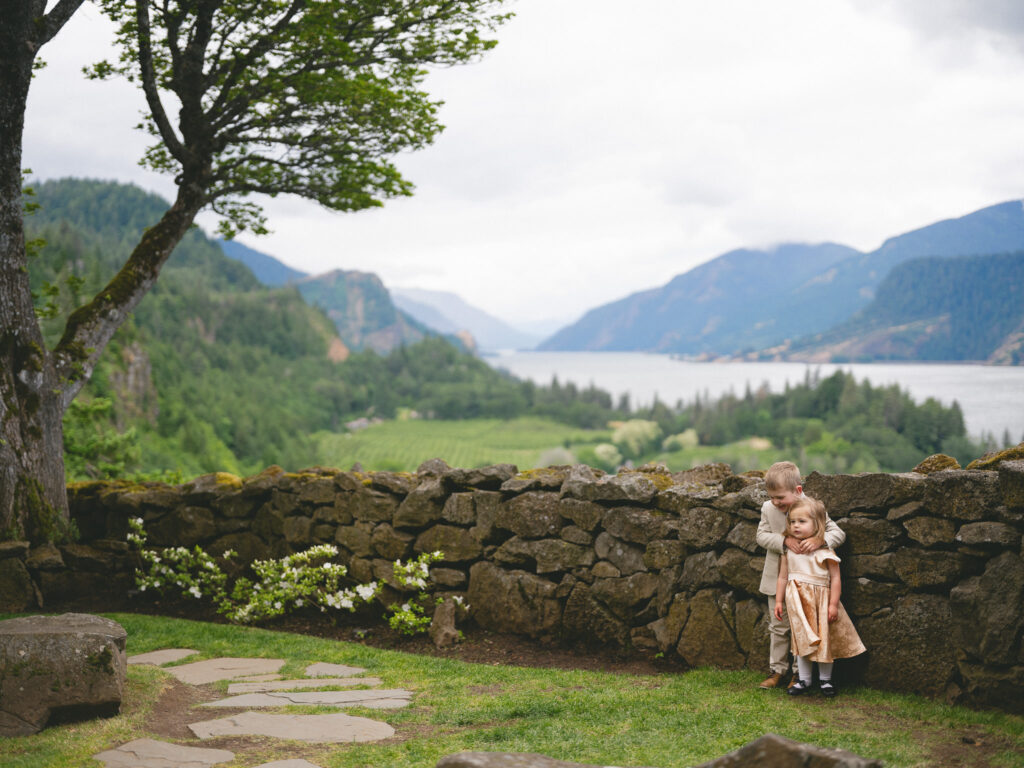 Children standing by a stone wall at the Griffin House, with sweeping views of the Columbia River Gorge behind them during a summer wedding.