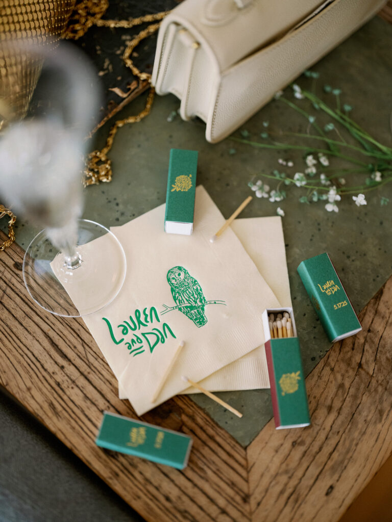 Flat lay of custom wedding matchbooks and cocktail napkins with an owl illustration and the couple’s names during a Griffin House wedding.