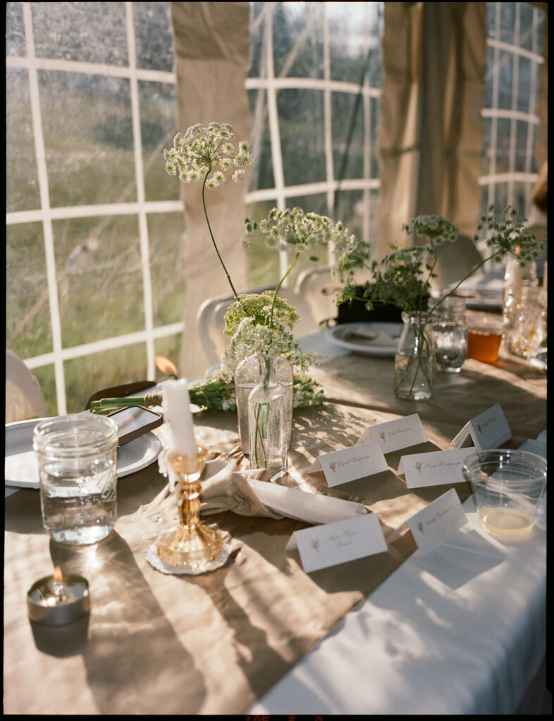 Reception table inside the wedding tent with Queen Anne’s lace in glass bottles, candlelight, mason jars, and handwritten place cards in soft evening light.