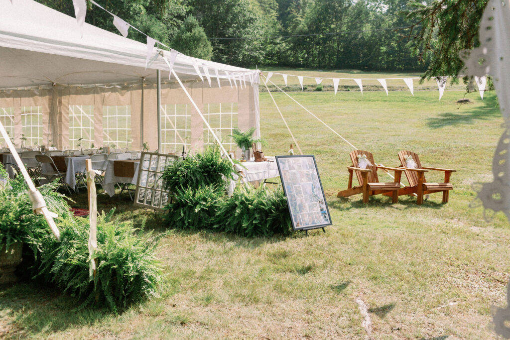 Outdoor wedding tent decorated with white bunting, lush ferns, framed photo displays, and two wooden Adirondack chairs on the lawn.