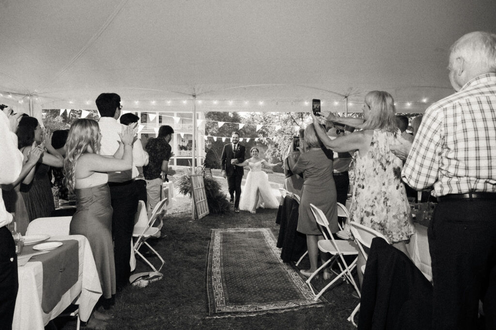 Bride and groom entering the tented reception to cheers and raised phones as guests stand to welcome them.
