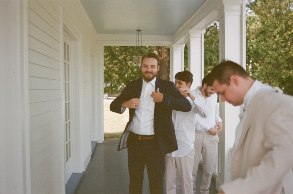 The groom puts on his suit jacket with help from his groomsmen on the porch of The White House on the Lake during the wedding morning.