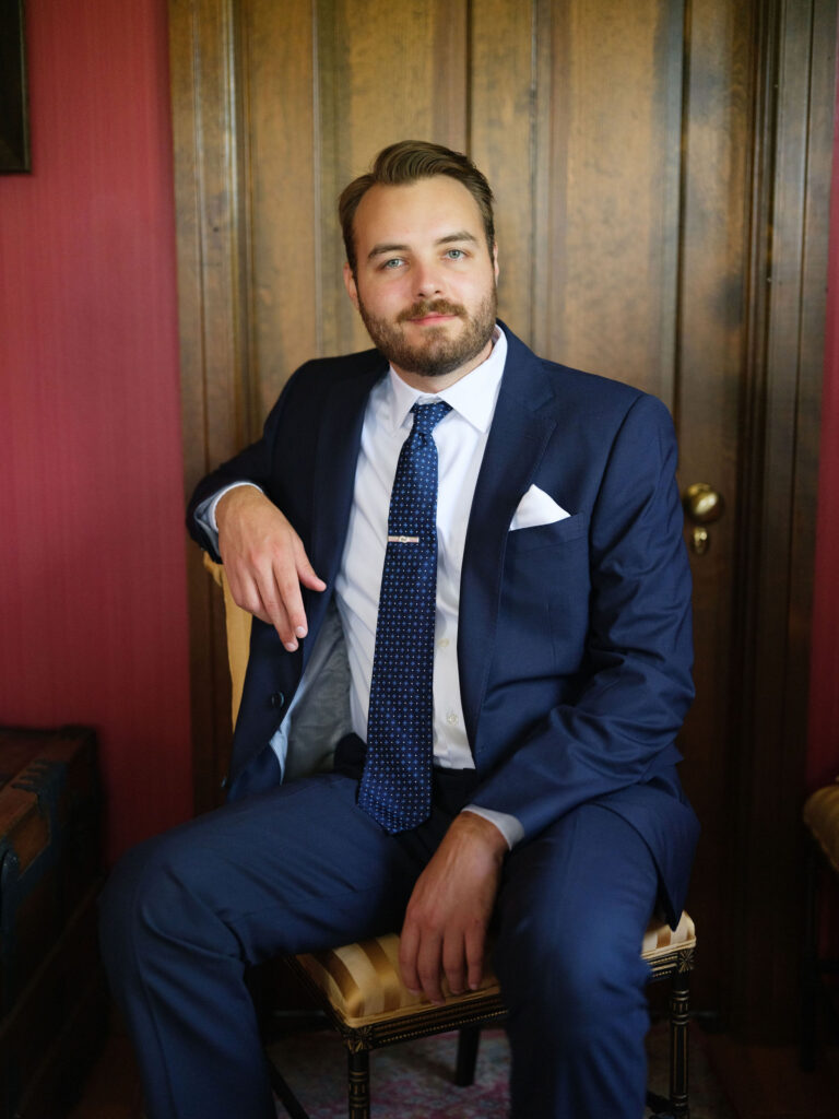 The groom sits in a vintage chair at The White House on the Lake, dressed in a navy suit and tie, surrounded by warm wood walls and classic décor.