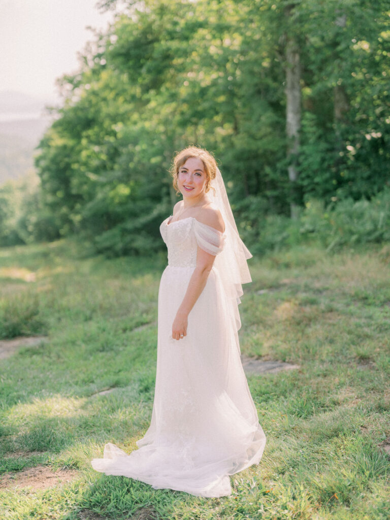 Bride standing alone on a sunlit hillside at Oak Mountain, wearing an off-the-shoulder gown and veil.