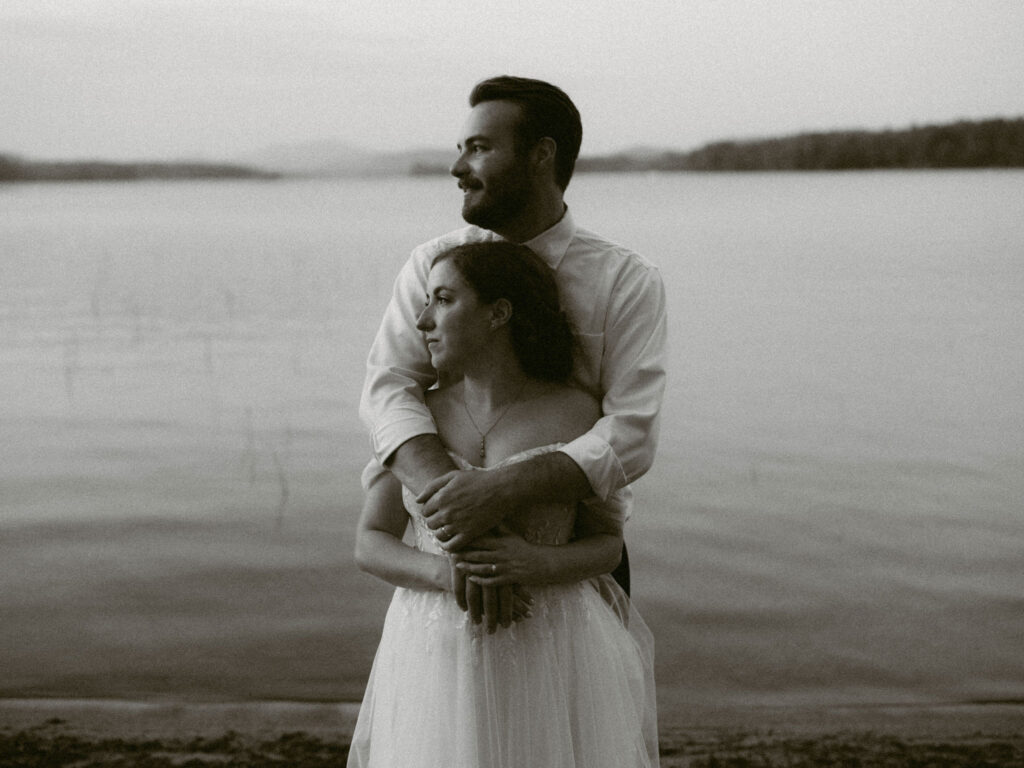 Bride and groom embracing by the lake at dusk, looking out toward the Adirondack mountains in soft evening light.