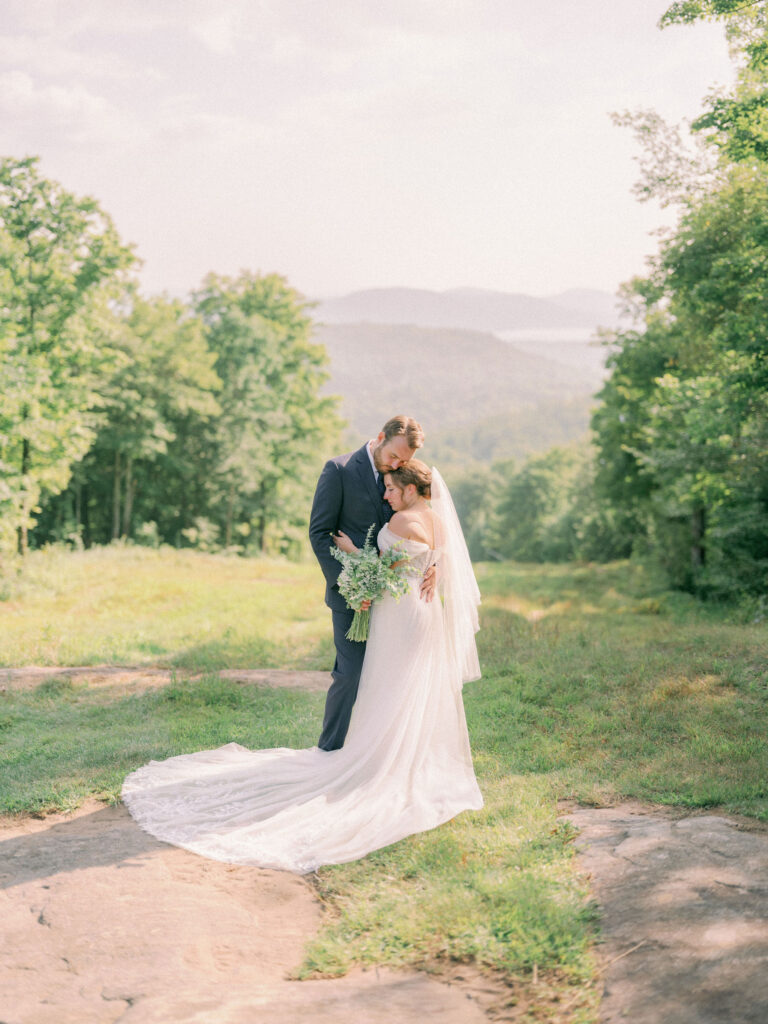 A bride and groom stand together on a sunlit overlook at Oak Mountain in Speculator, NY, embracing softly with rolling Adirondack hills in the background. The bride’s long, flowing gown trails over the stone path as the groom rests his forehead gently against hers, surrounded by summer greenery.