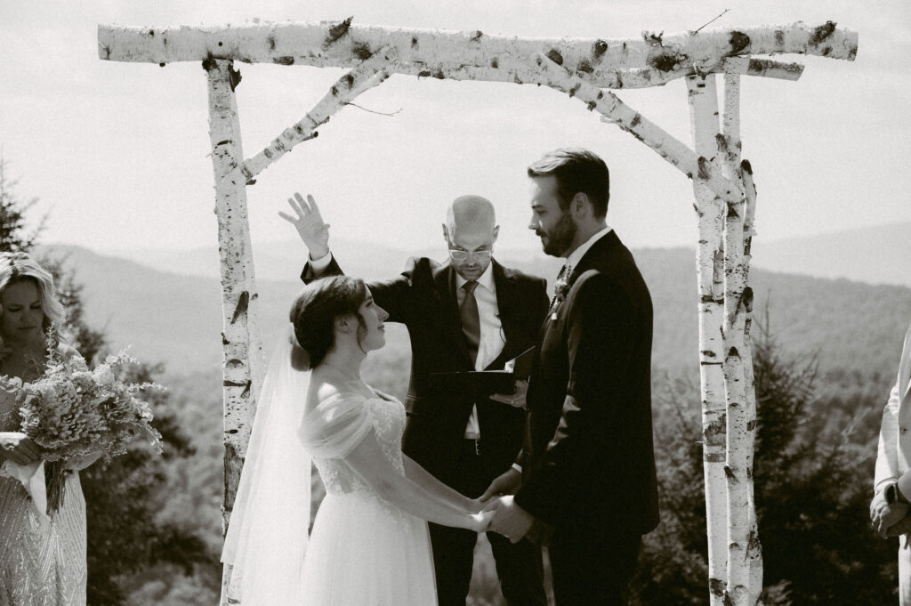Black-and-white photo of the officiant raising his hand in blessing as the bride and groom hold hands beneath the birch arch overlooking the Adirondack mountains.