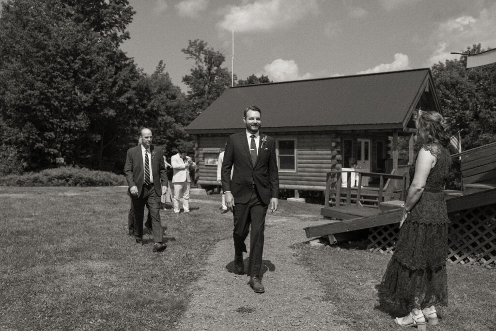 Black-and-white image of the groom walking toward the ceremony at Oak Mountain, smiling as guests look on.