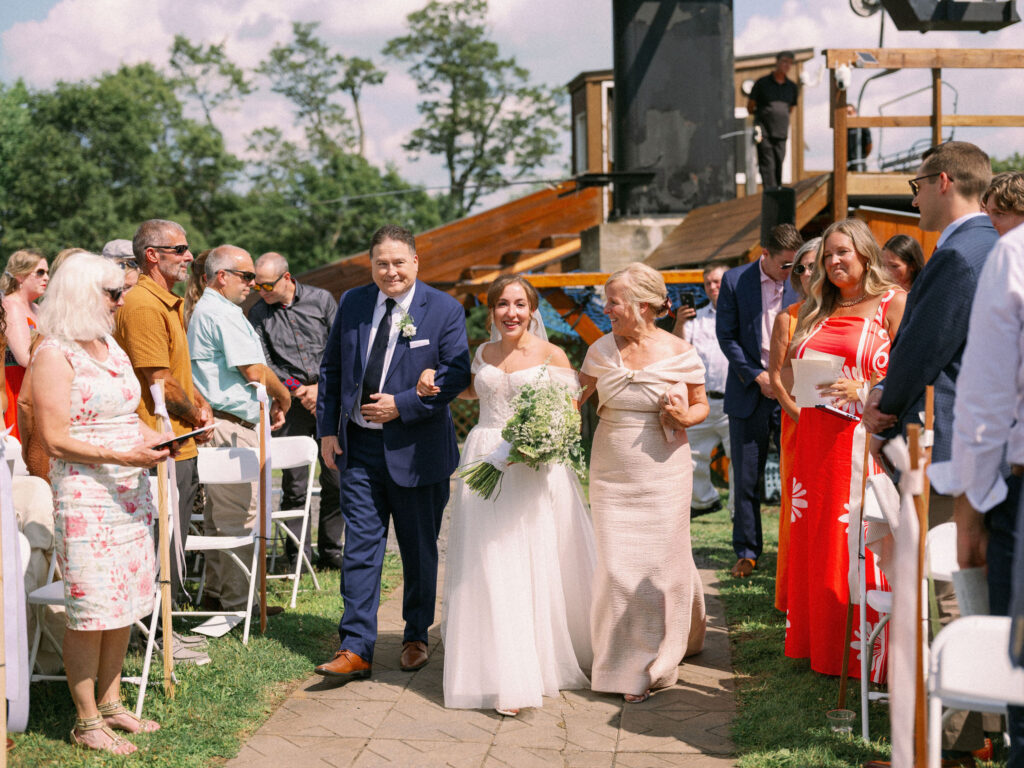 The bride and her parents walk together down the aisle toward the ceremony platform, surrounded by smiling guests in the summer sun.