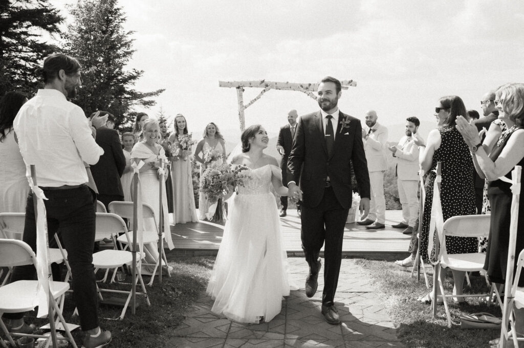 Black-and-white photo of the bride and groom walking back down the aisle together as guests stand and cheer at the conclusion of their Oak Mountain ceremony.