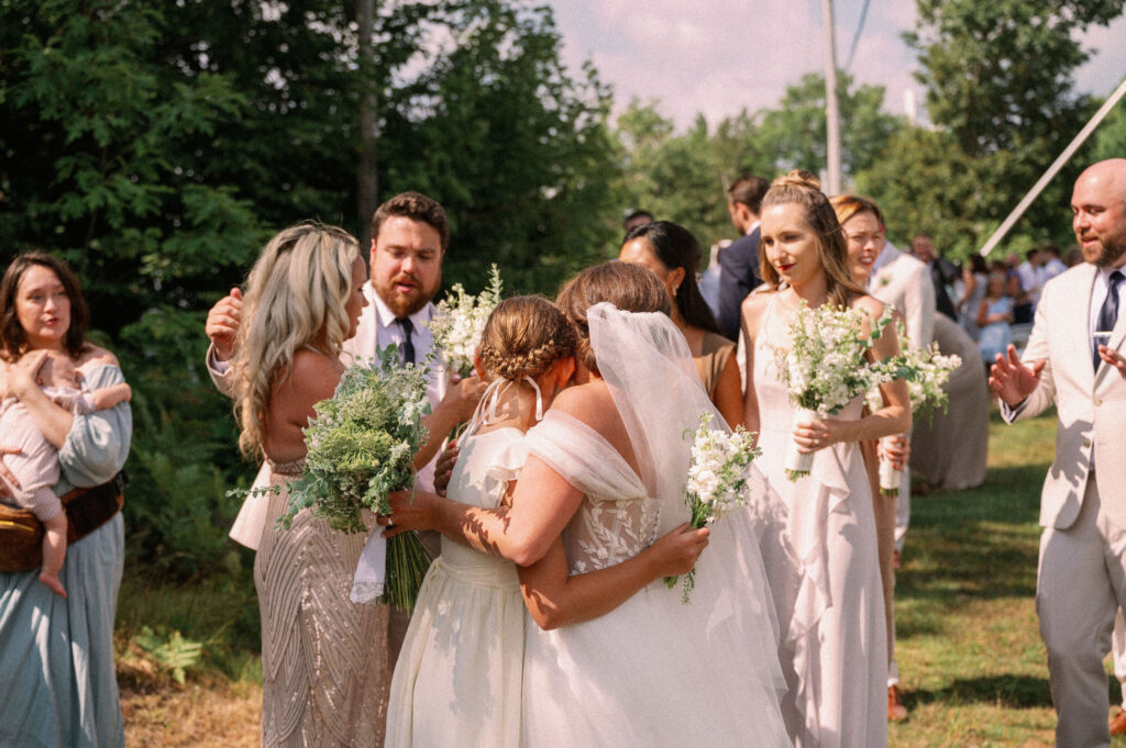 The bride embraces a bridesmaid in a warm, emotional moment after the ceremony, surrounded by family and friends with bouquets in hand.
