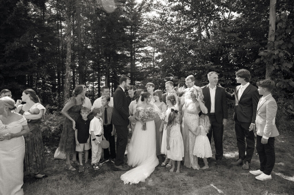 Black-and-white candid of the bride and groom surrounded by family members—children and adults—gathered together in the wooded clearing at Oak Mountain.