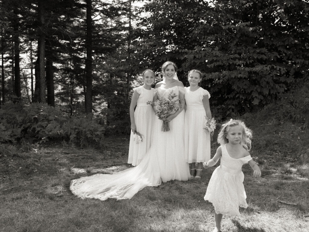 Black-and-white portrait of the bride standing with three young attendants near the woods at Oak Mountain, her long train sweeping across the grass as a flower girl runs playfully in the foreground.
