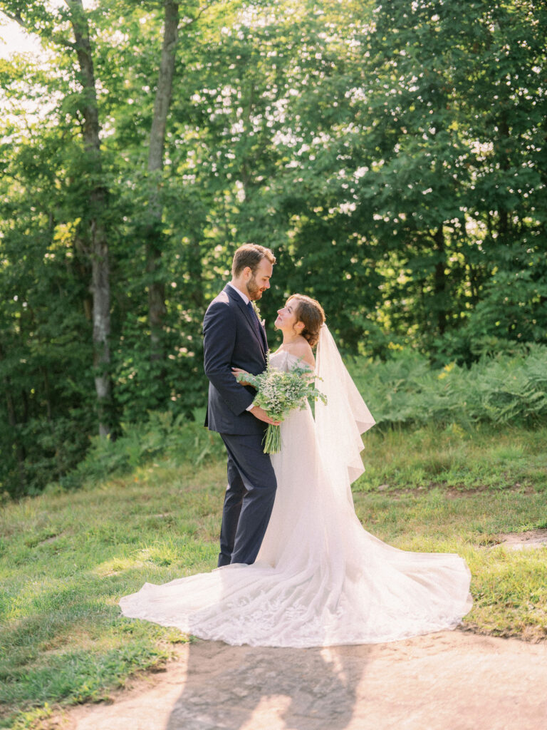 The bride and groom face each other in a sunlit clearing at Oak Mountain, surrounded by lush summer greenery.