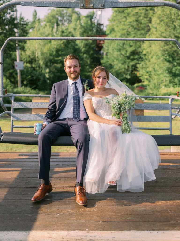 Bride and groom sitting together on the Oak Mountain ski lift platform, smiling in the late-day sun with her bouquet.