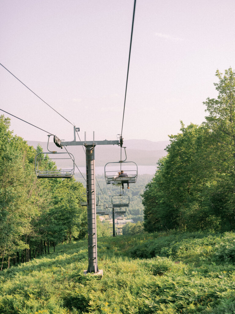 Chairlift ascending Oak Mountain through lush summer greenery with views of the lake and mountains.