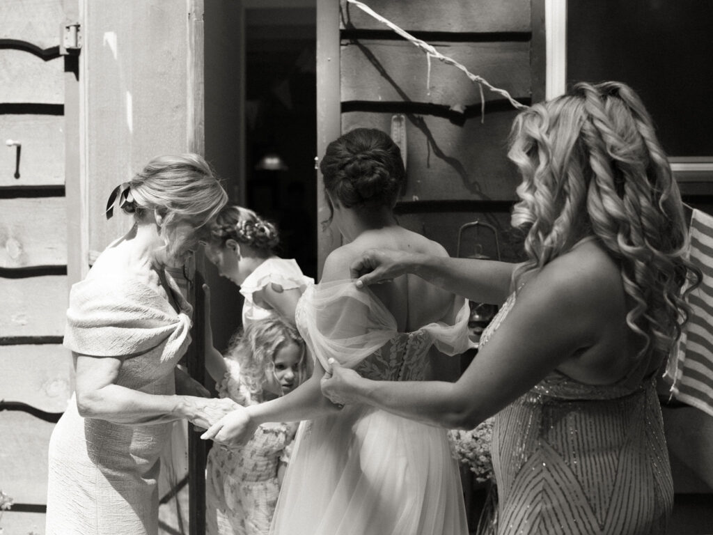Family members help adjust the bride’s gown and sleeves on the porch of the A-frame cabin, captured in a candid black-and-white moment.