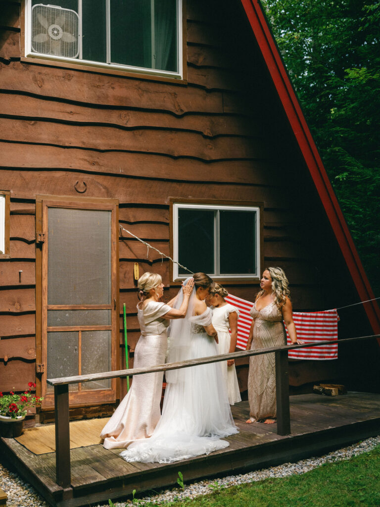 The bride stands on the porch of the A-frame cabin while her family helps secure her veil before the ceremony.