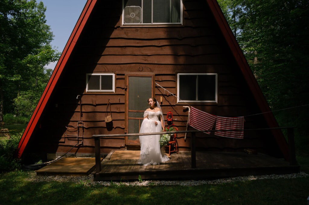 The bride stands on the porch of the rustic A-frame cabin in Speculator, NY, framed by dappled sunlight and surrounded by Adirondack forest.