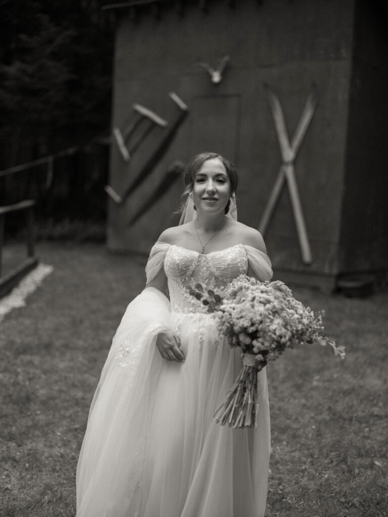 Black-and-white portrait of the bride holding her bouquet as she walks past a rustic shed decorated with old tools and wooden ladders.