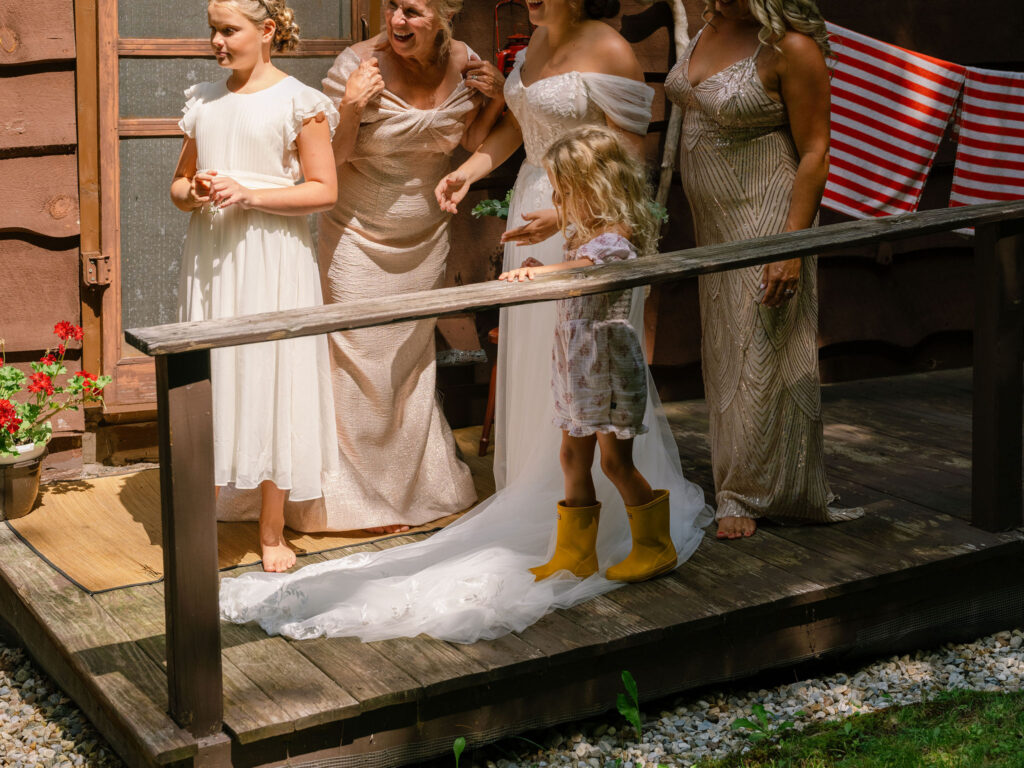 The bride and her family gather barefoot on the porch of the A-frame cabin, with sunlight falling across their dresses and a child in yellow boots at their feet.