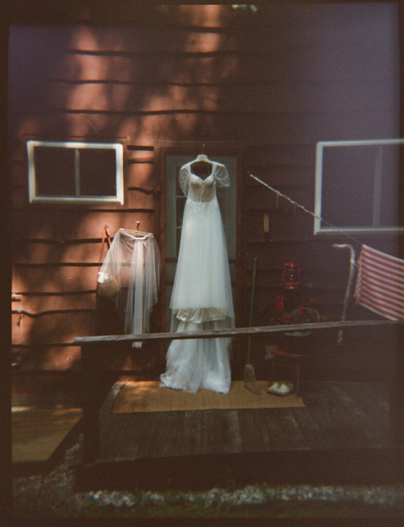 Film photograph of the wedding dress and veil hanging outside the rustic A-frame cabin, with dappled sunlight across the wooden siding.