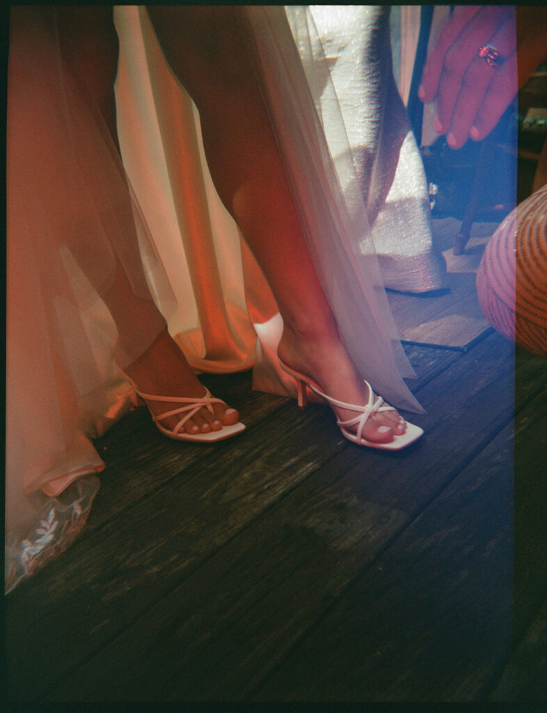 Film photograph of the bride slipping into her white heels, her gown sweeping softly across the wooden cabin floor.