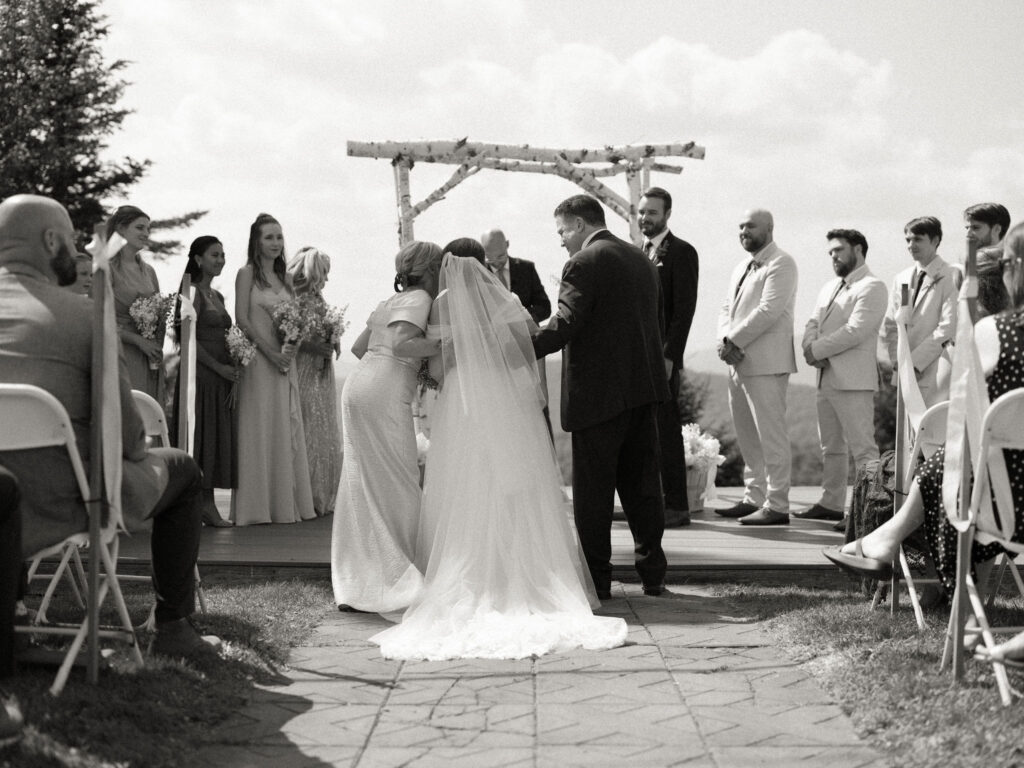 Black-and-white photo of the bride embracing her mother at the altar as the ceremony begins, with bridesmaids and groomsmen watching.